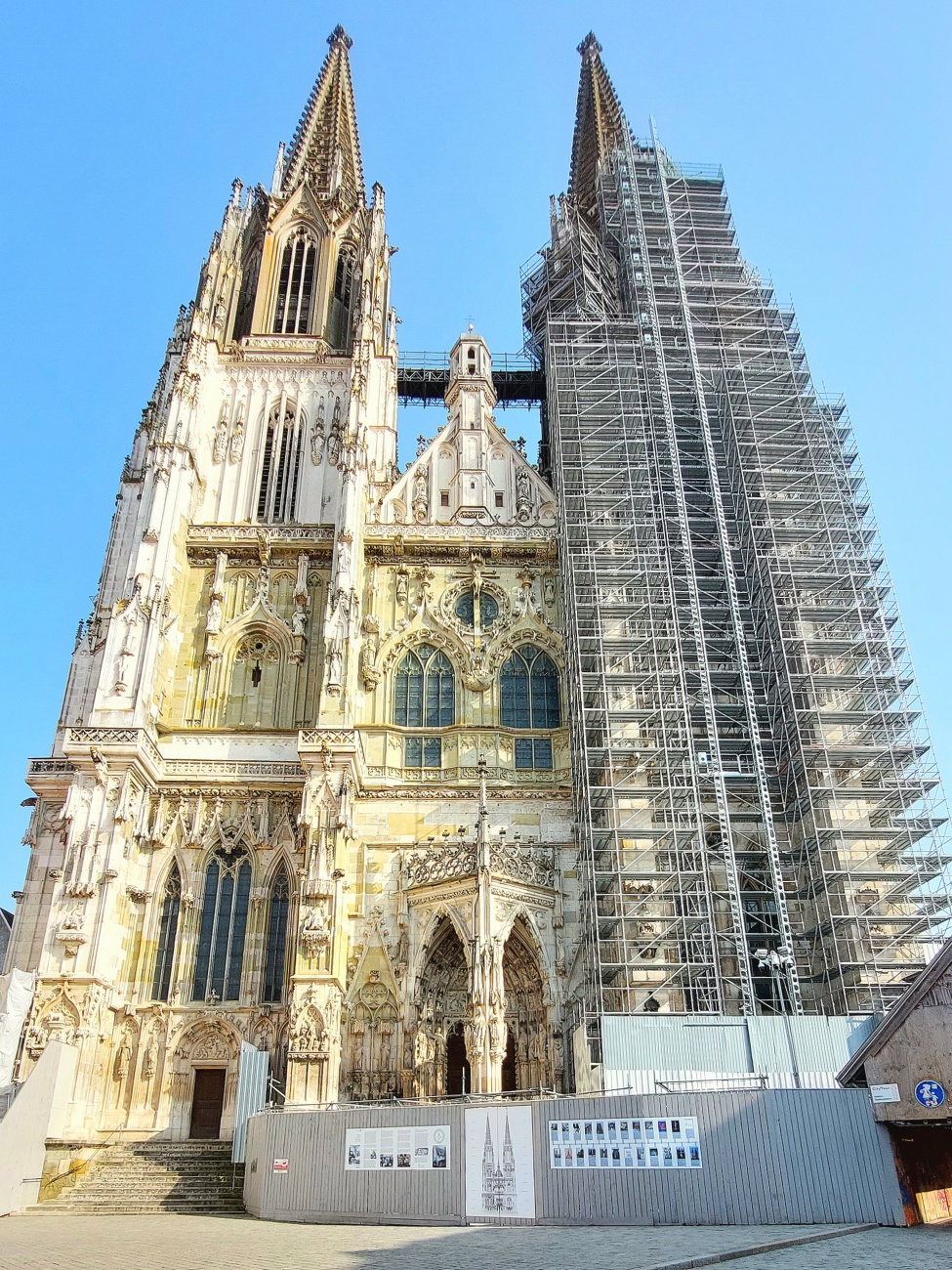 Auf diesem Foto ist der Regensburger Dom St. Peter zu sehen, teilweise eingerüstet als Teil umfangreicher Restaurierungs- und Erhaltungsarbeiten. 

Der markante Südturm ist aktuell von einem Baugerüst umgeben - ein Plan, der voraussichtlich bis 2029 andauern wird, sogar über das 750-jährige Dombaujubiläum hinaus, wenn alles nach Plan verläuft. Die Restaurierungsarbeiten am Südturm dienen vor allem dazu, belastete Pfeiler und kunstvolle Fassadenelemente fachgerecht zu sichern und zu erneuern, damit das Wahrzeichen Regensburgs auch für kommende Generationen erhalten bleibt. 

Der Regensburger Dom ist eine der bedeutendsten gotischen Kathedralen Deutschlands und zugleich die einzige gotische Kathedrale in Bayern. Seine Baugeschichte reicht zurück ins 13. Jahrhundert: Nach dem Brand des Vorgängerbaus begann 1275/76 der Neubau im gotischen Stil, der sich über mehrere Jahrhunderte erstreckte.  Die beeindruckenden Türme und Turmhelme, die heute das Stadtbild prägen, wurden erst zwischen 1859 und 1869 fertiggestellt. 

Das Bauwerk zeichnet sich besonders durch seine hohe gotische Architektur, filigrane Steinmetzarbeit und farbenprächtige Glasfenster aus dem 13. und 14. Jahrhundert aus, die zu den wertvollsten mittelalterlichen Glasmalereien im deutschen Raum gehören. 

Die Restaurierung des Doms ist ein fortlaufender Prozess: 

seit Jahrzehnten wird das Gebäude regelmäßig gepflegt, gereinigt und instand gesetzt, da Witterung, Alter und Schadstoffe dem Kalkstein zusetzen. Die sogenannte Dombauhütte, eine spezialisierte Werkstatt für Steinmetze und Restauratoren, arbeitet nahezu ununterbrochen am Erhalt dieses einzigartigen Bauwerks - weshalb der Dom oft eingerüstet ist.