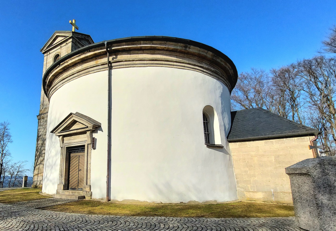 Die Wallfahrtskirche auf dem Armesberg bei schönem Frühlingswetter

Bei schönem Frühlingswetter zeigt sich die Wallfahrtskirche zur Heiligsten Dreifaltigkeit auf dem Armesberg von ihrer eindrucksvollsten Seite. Der Armesberg erhebt sich bis auf 731 Meter über Normalnull und ist ein markanter Basaltkegel im nördlichen Teil der Oberpfalz. Als ehemaliger Vulkanschlot ragt er deutlich aus der umgebenden Landschaft hervor und bietet eine weite Aussicht über das Fichtelgebirge und das Umland.

Auf seinem Gipfel steht die Kirche mit ihrem charakteristischen runden Baukörper. Die hell verputzten Mauern leuchten im Sonnenlicht, während sich darüber der kleine Turm mit Kreuz gegen den blauen Himmel abzeichnet. Das schlichte, aber kraftvolle Erscheinungsbild des Gotteshauses fügt sich harmonisch in die natürliche Umgebung ein. Gerade im Frühling, wenn die Bäume austreiben und die Wiesen wieder grün werden, entsteht eine besonders freundliche und zugleich feierliche Atmosphäre.

Die Geschichte des Wallfahrtsortes reicht bis ins 17. Jahrhundert zurück. In den Jahren 1677 und 1678 wurde auf dem Berg eine erste Kapelle errichtet. Die heutige Kirche entstand später. Der Bau begann im Jahr 1822 und die Weihe erfolgte 1836. Seitdem ist der Armesberg ein bedeutender Ort des Glaubens und der regionalen Wallfahrt. Ein Kreuzweg führt hinauf zur Kirche und unterstreicht die religiöse Bedeutung des Berges.

Wer an einem klaren Frühlingstag den Gipfel erreicht, spürt die besondere Verbindung von Natur, Geschichte und Spiritualität. Das Licht, die frische Luft und die weite Aussicht verleihen dem Ort eine ruhige und zugleich erhabene Stimmung. Die Kirche auf dem Armesberg ist damit nicht nur ein historisches Bauwerk, sondern auch ein lebendiger Treffpunkt für Pilger, Wanderer und Besucher, die hier oben einen Moment der Stille suchen.