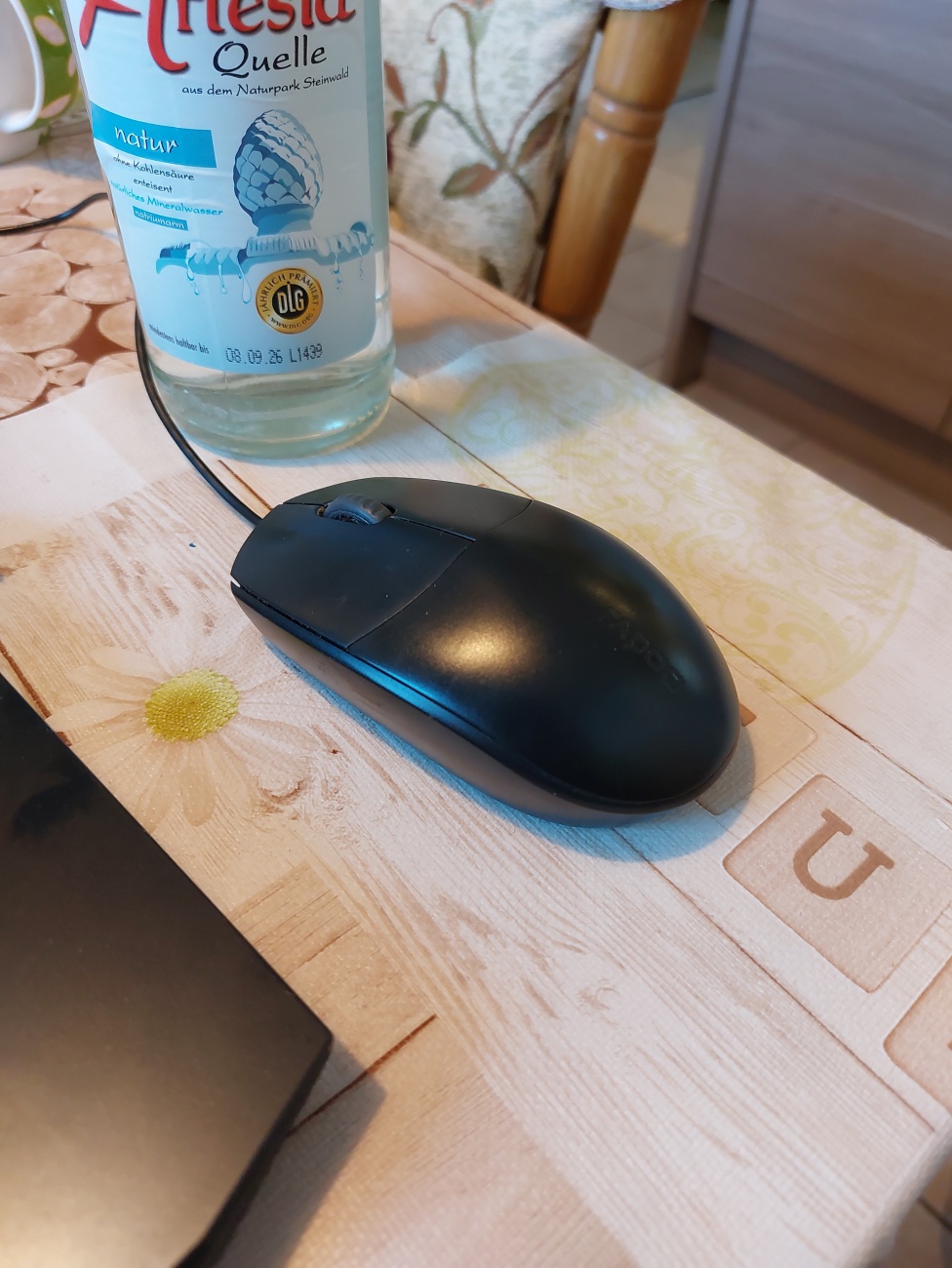 Foto: Martin Zehrer - Das Foto zeigt eine Computermaus, die auf einem holzähnlichen Untergrund liegt, neben einer Flasche Mineralwasser. Die Maus besteht aus Kunststoff und besitzt eine ergono 
