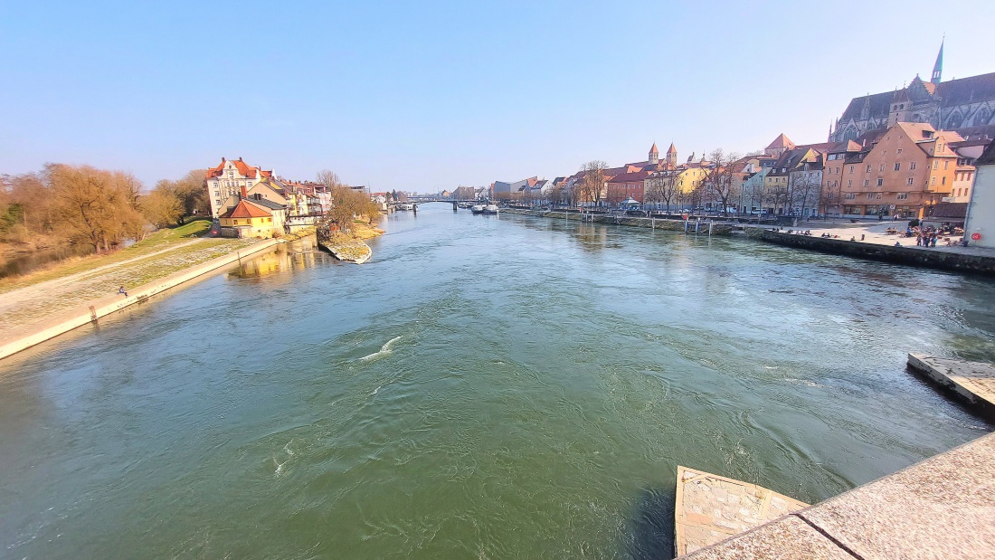 Frühlingstag in Regensburg - Blick von der Steinernen Brücke

Am 9. März 2026 zeigt sich Regensburg von seiner schönsten Seite. Bei strahlendem Sonnenschein und milden 15 Grad eröffnet sich von der Steinernen Brücke ein beeindruckender Blick über die Donau auf die historische Altstadt. Im Hintergrund erhebt sich majestätisch der Regensburger Dom und prägt die Silhouette der Stadt.

Unterhalb der Brücke fließt die Donau ruhig dahin, während sich am Ufer bereits die ersten Menschen des Jahres im grünen Uferstreifen niederlassen, um die warmen Sonnenstrahlen zu genießen. Ein wunderschöner Frühlingstag, der die besondere Atmosphäre dieser geschichtsträchtigen Stadt spürbar macht.