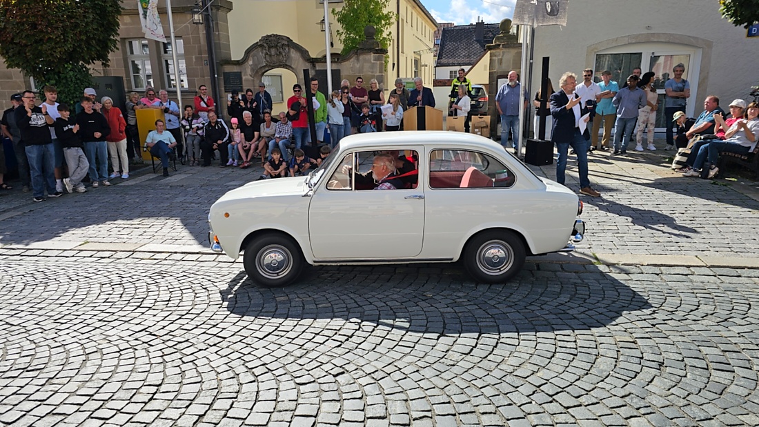 Jede Menge automobile Schätze auf dem Kemnather Stadtplatz

Ein echtes Paradies für Oldtimerfreunde gab es am Samstag beim großen Oldtimertreffen in Kemnath zu erleben. Ab 10 Uhr gehörte der wunderschöne historische Stadtplatz den automobilen Schätzen vergangener Jahrzehnte. Bei bestem Oldtimerwetter präsentierten sich die liebevoll gepflegten Fahrzeuge vor einer traumhaften Kulisse – und natürlich ließen sich das auch jede Menge Zuschauer nicht entgehen.

Dicht an dicht standen die Besucher entlang des Stadtplatzes, als ein Klassiker nach dem anderen zur Fahrzeugvorstellung vorfuhr. Dabei gab es nicht nur jede Menge Chrom, glänzenden Lack und den unverwechselbaren Klang alter Motoren zu bestaunen, sondern auch viel Wissenswertes zu hören.

Wie jedes Jahr hatte Elmar Högl das Mikrofon fest im Griff. Souverän und mit enormem Fachwissen stellte er die einzelnen Fahrzeuge vor, erzählte von Baujahren, Motorisierung und technischen Besonderheiten und hatte dabei immer wieder interessante Details zu den Oldtimern parat.

Und eigentlich ist Elmar Högl aus dem Kemnather Oldtimertreffen kaum wegzudenken. Er ist nicht nur die bekannte Stimme am Mikrofon, sondern zugleich Hauptorganisator und so etwas wie das „Herz“ des Treffens. Gemeinsam mit dem KEM-Verband und der Stadt Kemnath sorgt er dafür, dass die Freunde historischer Fahrzeuge Jahr für Jahr in Kemnath zusammenkommen.

Zur Fahrzeugvorstellung gehörte natürlich auch die Übergabe der Urkunden. Diese Aufgabe übernahm Hermann Schraml, unterstützt von Stefan Wöhrl.

Und besser hätte es an diesem Vormittag eigentlich kaum passen können: herrliches Wetter, ein voller Stadtplatz, viele begeisterte Zuschauer und mittendrin jede Menge automobile Zeitgeschichte auf zwei und vier Rädern.

Nach der Vorstellung blieb noch genügend Zeit, die Oldtimer aus nächster Nähe zu betrachten, mit den Besitzern ins Gespräch zu kommen und natürlich jede Menge Fotos zu machen.

Am Nachmittag wartete dann bereits der nächste Höhepunkt auf die Teilnehmer. Um 13:15 Uhr machten sich die historischen Fahrzeuge zur gemeinsamen Ausfahrt auf den Weg. Über Berndorf, Haidenaab und Weidenberg führte die Tour unter anderem nach Goldkronach, wo eine längere Rast eingelegt wurde, bevor es schließlich wieder zurück nach Kemnath ging.

Und jetzt viel Spaß beim Durchklicken durch unsere Bilder vom Kemnather Oldtimertreffen 2026!