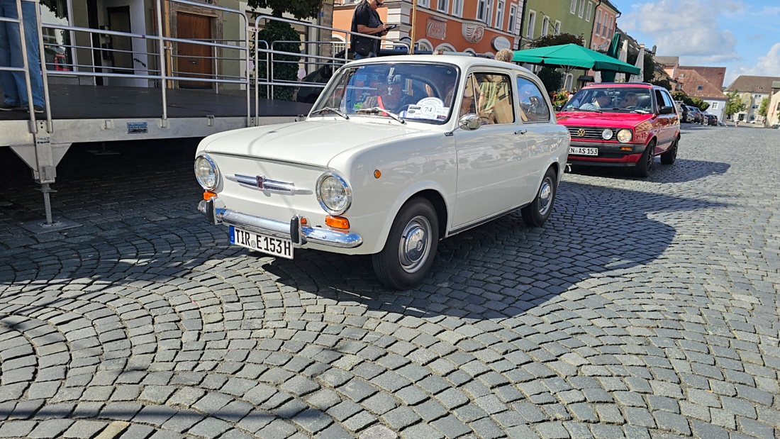 Jede Menge automobile Schätze auf dem Kemnather Stadtplatz

Ein echtes Paradies für Oldtimerfreunde gab es am Samstag beim großen Oldtimertreffen in Kemnath zu erleben. Ab 10 Uhr gehörte der wunderschöne historische Stadtplatz den automobilen Schätzen vergangener Jahrzehnte. Bei bestem Oldtimerwetter präsentierten sich die liebevoll gepflegten Fahrzeuge vor einer traumhaften Kulisse – und natürlich ließen sich das auch jede Menge Zuschauer nicht entgehen.

Dicht an dicht standen die Besucher entlang des Stadtplatzes, als ein Klassiker nach dem anderen zur Fahrzeugvorstellung vorfuhr. Dabei gab es nicht nur jede Menge Chrom, glänzenden Lack und den unverwechselbaren Klang alter Motoren zu bestaunen, sondern auch viel Wissenswertes zu hören.

Wie jedes Jahr hatte Elmar Högl das Mikrofon fest im Griff. Souverän und mit enormem Fachwissen stellte er die einzelnen Fahrzeuge vor, erzählte von Baujahren, Motorisierung und technischen Besonderheiten und hatte dabei immer wieder interessante Details zu den Oldtimern parat.

Und eigentlich ist Elmar Högl aus dem Kemnather Oldtimertreffen kaum wegzudenken. Er ist nicht nur die bekannte Stimme am Mikrofon, sondern zugleich Hauptorganisator und so etwas wie das „Herz“ des Treffens. Gemeinsam mit dem KEM-Verband und der Stadt Kemnath sorgt er dafür, dass die Freunde historischer Fahrzeuge Jahr für Jahr in Kemnath zusammenkommen.

Zur Fahrzeugvorstellung gehörte natürlich auch die Übergabe der Urkunden. Diese Aufgabe übernahm Hermann Schraml, unterstützt von Stefan Wöhrl.

Und besser hätte es an diesem Vormittag eigentlich kaum passen können: herrliches Wetter, ein voller Stadtplatz, viele begeisterte Zuschauer und mittendrin jede Menge automobile Zeitgeschichte auf zwei und vier Rädern.

Nach der Vorstellung blieb noch genügend Zeit, die Oldtimer aus nächster Nähe zu betrachten, mit den Besitzern ins Gespräch zu kommen und natürlich jede Menge Fotos zu machen.

Am Nachmittag wartete dann bereits der nächste Höhepunkt auf die Teilnehmer. Um 13:15 Uhr machten sich die historischen Fahrzeuge zur gemeinsamen Ausfahrt auf den Weg. Über Berndorf, Haidenaab und Weidenberg führte die Tour unter anderem nach Goldkronach, wo eine längere Rast eingelegt wurde, bevor es schließlich wieder zurück nach Kemnath ging.

Und jetzt viel Spaß beim Durchklicken durch unsere Bilder vom Kemnather Oldtimertreffen 2026!