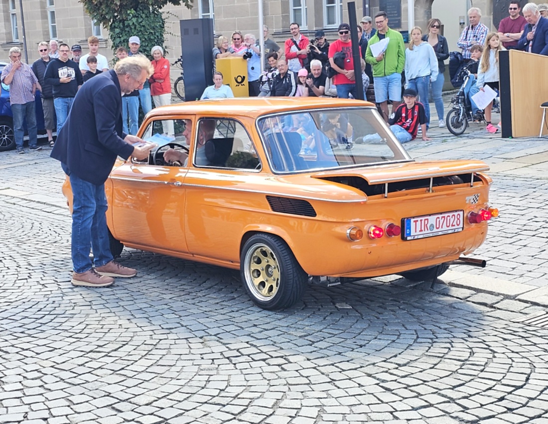 Jede Menge automobile Schätze auf dem Kemnather Stadtplatz

Ein echtes Paradies für Oldtimerfreunde gab es am Samstag beim großen Oldtimertreffen in Kemnath zu erleben. Ab 10 Uhr gehörte der wunderschöne historische Stadtplatz den automobilen Schätzen vergangener Jahrzehnte. Bei bestem Oldtimerwetter präsentierten sich die liebevoll gepflegten Fahrzeuge vor einer traumhaften Kulisse – und natürlich ließen sich das auch jede Menge Zuschauer nicht entgehen.

Dicht an dicht standen die Besucher entlang des Stadtplatzes, als ein Klassiker nach dem anderen zur Fahrzeugvorstellung vorfuhr. Dabei gab es nicht nur jede Menge Chrom, glänzenden Lack und den unverwechselbaren Klang alter Motoren zu bestaunen, sondern auch viel Wissenswertes zu hören.

Wie jedes Jahr hatte Elmar Högl das Mikrofon fest im Griff. Souverän und mit enormem Fachwissen stellte er die einzelnen Fahrzeuge vor, erzählte von Baujahren, Motorisierung und technischen Besonderheiten und hatte dabei immer wieder interessante Details zu den Oldtimern parat.

Und eigentlich ist Elmar Högl aus dem Kemnather Oldtimertreffen kaum wegzudenken. Er ist nicht nur die bekannte Stimme am Mikrofon, sondern zugleich Hauptorganisator und so etwas wie das „Herz“ des Treffens. Gemeinsam mit dem KEM-Verband und der Stadt Kemnath sorgt er dafür, dass die Freunde historischer Fahrzeuge Jahr für Jahr in Kemnath zusammenkommen.

Zur Fahrzeugvorstellung gehörte natürlich auch die Übergabe der Urkunden. Diese Aufgabe übernahm Hermann Schraml, unterstützt von Stefan Wöhrl.

Und besser hätte es an diesem Vormittag eigentlich kaum passen können: herrliches Wetter, ein voller Stadtplatz, viele begeisterte Zuschauer und mittendrin jede Menge automobile Zeitgeschichte auf zwei und vier Rädern.

Nach der Vorstellung blieb noch genügend Zeit, die Oldtimer aus nächster Nähe zu betrachten, mit den Besitzern ins Gespräch zu kommen und natürlich jede Menge Fotos zu machen.

Am Nachmittag wartete dann bereits der nächste Höhepunkt auf die Teilnehmer. Um 13:15 Uhr machten sich die historischen Fahrzeuge zur gemeinsamen Ausfahrt auf den Weg. Über Berndorf, Haidenaab und Weidenberg führte die Tour unter anderem nach Goldkronach, wo eine längere Rast eingelegt wurde, bevor es schließlich wieder zurück nach Kemnath ging.

Und jetzt viel Spaß beim Durchklicken durch unsere Bilder vom Kemnather Oldtimertreffen 2026!