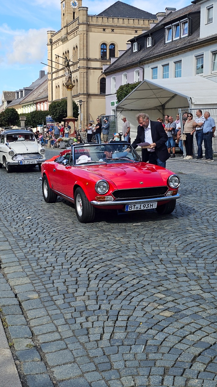 Jede Menge automobile Schätze auf dem Kemnather Stadtplatz

Ein echtes Paradies für Oldtimerfreunde gab es am Samstag beim großen Oldtimertreffen in Kemnath zu erleben. Ab 10 Uhr gehörte der wunderschöne historische Stadtplatz den automobilen Schätzen vergangener Jahrzehnte. Bei bestem Oldtimerwetter präsentierten sich die liebevoll gepflegten Fahrzeuge vor einer traumhaften Kulisse – und natürlich ließen sich das auch jede Menge Zuschauer nicht entgehen.

Dicht an dicht standen die Besucher entlang des Stadtplatzes, als ein Klassiker nach dem anderen zur Fahrzeugvorstellung vorfuhr. Dabei gab es nicht nur jede Menge Chrom, glänzenden Lack und den unverwechselbaren Klang alter Motoren zu bestaunen, sondern auch viel Wissenswertes zu hören.

Wie jedes Jahr hatte Elmar Högl das Mikrofon fest im Griff. Souverän und mit enormem Fachwissen stellte er die einzelnen Fahrzeuge vor, erzählte von Baujahren, Motorisierung und technischen Besonderheiten und hatte dabei immer wieder interessante Details zu den Oldtimern parat.

Und eigentlich ist Elmar Högl aus dem Kemnather Oldtimertreffen kaum wegzudenken. Er ist nicht nur die bekannte Stimme am Mikrofon, sondern zugleich Hauptorganisator und so etwas wie das „Herz“ des Treffens. Gemeinsam mit dem KEM-Verband und der Stadt Kemnath sorgt er dafür, dass die Freunde historischer Fahrzeuge Jahr für Jahr in Kemnath zusammenkommen.

Zur Fahrzeugvorstellung gehörte natürlich auch die Übergabe der Urkunden. Diese Aufgabe übernahm Hermann Schraml, unterstützt von Stefan Wöhrl.

Und besser hätte es an diesem Vormittag eigentlich kaum passen können: herrliches Wetter, ein voller Stadtplatz, viele begeisterte Zuschauer und mittendrin jede Menge automobile Zeitgeschichte auf zwei und vier Rädern.

Nach der Vorstellung blieb noch genügend Zeit, die Oldtimer aus nächster Nähe zu betrachten, mit den Besitzern ins Gespräch zu kommen und natürlich jede Menge Fotos zu machen.

Am Nachmittag wartete dann bereits der nächste Höhepunkt auf die Teilnehmer. Um 13:15 Uhr machten sich die historischen Fahrzeuge zur gemeinsamen Ausfahrt auf den Weg. Über Berndorf, Haidenaab und Weidenberg führte die Tour unter anderem nach Goldkronach, wo eine längere Rast eingelegt wurde, bevor es schließlich wieder zurück nach Kemnath ging.

Und jetzt viel Spaß beim Durchklicken durch unsere Bilder vom Kemnather Oldtimertreffen 2026!