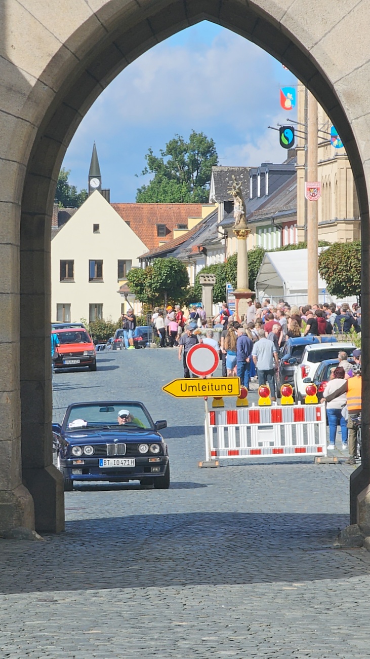 Jede Menge automobile Schätze auf dem Kemnather Stadtplatz

Ein echtes Paradies für Oldtimerfreunde gab es am Samstag beim großen Oldtimertreffen in Kemnath zu erleben. Ab 10 Uhr gehörte der wunderschöne historische Stadtplatz den automobilen Schätzen vergangener Jahrzehnte. Bei bestem Oldtimerwetter präsentierten sich die liebevoll gepflegten Fahrzeuge vor einer traumhaften Kulisse – und natürlich ließen sich das auch jede Menge Zuschauer nicht entgehen.

Dicht an dicht standen die Besucher entlang des Stadtplatzes, als ein Klassiker nach dem anderen zur Fahrzeugvorstellung vorfuhr. Dabei gab es nicht nur jede Menge Chrom, glänzenden Lack und den unverwechselbaren Klang alter Motoren zu bestaunen, sondern auch viel Wissenswertes zu hören.

Wie jedes Jahr hatte Elmar Högl das Mikrofon fest im Griff. Souverän und mit enormem Fachwissen stellte er die einzelnen Fahrzeuge vor, erzählte von Baujahren, Motorisierung und technischen Besonderheiten und hatte dabei immer wieder interessante Details zu den Oldtimern parat.

Und eigentlich ist Elmar Högl aus dem Kemnather Oldtimertreffen kaum wegzudenken. Er ist nicht nur die bekannte Stimme am Mikrofon, sondern zugleich Hauptorganisator und so etwas wie das „Herz“ des Treffens. Gemeinsam mit dem KEM-Verband und der Stadt Kemnath sorgt er dafür, dass die Freunde historischer Fahrzeuge Jahr für Jahr in Kemnath zusammenkommen.

Zur Fahrzeugvorstellung gehörte natürlich auch die Übergabe der Urkunden. Diese Aufgabe übernahm Hermann Schraml, unterstützt von Stefan Wöhrl.

Und besser hätte es an diesem Vormittag eigentlich kaum passen können: herrliches Wetter, ein voller Stadtplatz, viele begeisterte Zuschauer und mittendrin jede Menge automobile Zeitgeschichte auf zwei und vier Rädern.

Nach der Vorstellung blieb noch genügend Zeit, die Oldtimer aus nächster Nähe zu betrachten, mit den Besitzern ins Gespräch zu kommen und natürlich jede Menge Fotos zu machen.

Am Nachmittag wartete dann bereits der nächste Höhepunkt auf die Teilnehmer. Um 13:15 Uhr machten sich die historischen Fahrzeuge zur gemeinsamen Ausfahrt auf den Weg. Über Berndorf, Haidenaab und Weidenberg führte die Tour unter anderem nach Goldkronach, wo eine längere Rast eingelegt wurde, bevor es schließlich wieder zurück nach Kemnath ging.

Und jetzt viel Spaß beim Durchklicken durch unsere Bilder vom Kemnather Oldtimertreffen 2026!