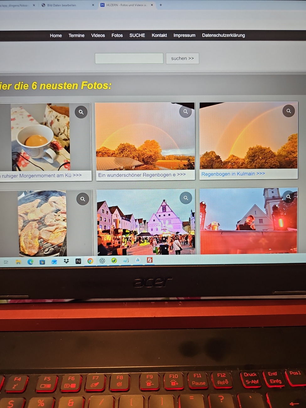 In diesem Moment entfaltet sich auf dem Laptopbildschirm ein charmantes Foto-Mosaik aus regionalem Alltag und visueller Ruhe: Eine Software-Oberfläche mit graubraunen Menüleisten für „Home“, „Termine“, „Videos“, „Fotos“, „Suche“, „Kontakt“, „Impressum“ und „Datenschutzerklärung“ vermittelt einen minimalistischen, funktionalen Stil, und darunter lädt die Seite augenblicklich mit dem Hinweis auf „Hier die 6 neuesten Fotos:“ zum digitalen Stöbern ein. Die abgebildeten Vorschaubilder zeigen unterschiedliche Motive wie einen ruhigen Morgenkaffee in einer Tasse, einen bunten Regenbogen über herbstlich verfärbtem Laub und ein beleuchtetes Stadtgeschehen mit Fachwerkhäusern – eine kontrastreiche Mischung aus Natur, Genuss und regionaler Atmosphäre, die auf eine vielseitige Fotodatenbank regionaler Momente schließen lässt. Im Vordergrund vibrieren die Tasten eines beleuchteten Acer-Laptops, dessen angedeutete Ränder Bodenhaftung verleihen und das Foto in einen spannenden Kontrast von digitaler Technik und analoger Archivierung setzen. Das Bild fängt den unspektakulären Reiz des Alltags ein, lädt neugierig ein, weiterzuklicken und mehr von diesen lebendigen, regional verankerten Eindrücken zu entdecken — ein stimmiger Einstieg für eine Plattform, die kleine Geschichten groß und heimische Augenblicke sichtbar macht.