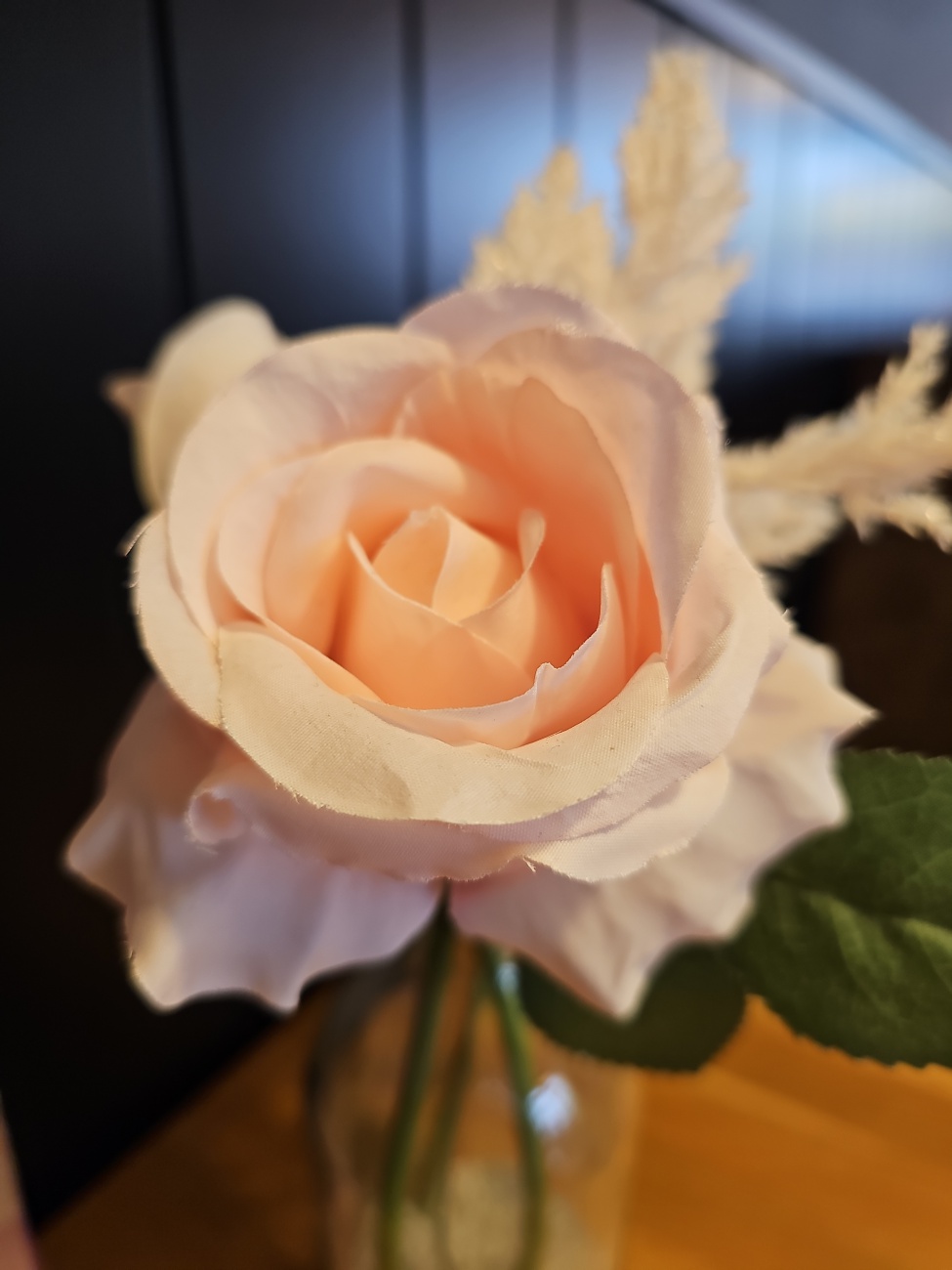 Die zarte, blassrosafarbene Rose mit ihren samtig wirkenden, fein geschwungenen Blütenblättern vermittelt eine bemerkenswerte Ruhe – als stamme sie nicht aus einem Garten, sondern aus einem dekorativen Arrangement. Die klar konturierten Ränder der Blätter und die unnatürliche Perfektion der Schichtung deuten auf eine hochwertige Kunstblume hin, möglicherweise aus Textil oder Schaumstoff gefertigt, die echte Rosen dekorativ imitiert. Hochwertige künstliche Rosen dieser Art werden oft aus Polyester- oder Latexmaterial hergestellt, um eine realistische Haptik und matte Oberfläche zu erzielen, wie bei sogenannten „Real-Touch“- oder seidenähnlichen Varianten üblich ist. In diesem stillen Bild nimmt die Rose die Kulisse nur zurückhaltend wahr, während ihr samtiger Farbverlauf und die klare Form die Blicke souverän auf sich ziehen. Sie wirkt wie ein stummer Begleiter im Alltag, der klassisch-elegante Schönheit in eine Umgebung bringt, ohne je zu welken oder Pflege zu verlangen. Ihre Beständigkeit macht sie zur idealen Dekoration für Wohnräume, auf dem Fensterbrett oder als dauerhafte Tischschmuckidee – ästhetisch, unvergänglich und ganz ohne Pflegeaufwand.