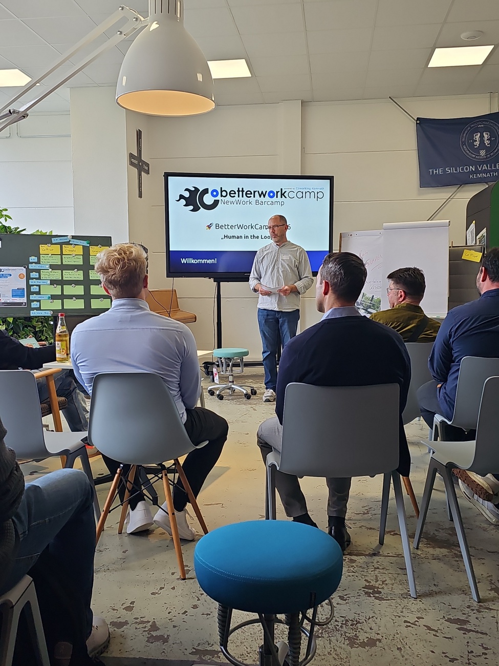 Ein Moderator, Thomas Völkl, eröffnet das BetterWorkCamp unter dem Motto ..Human in the Loop“ mit einem offenen Barcamp, bei dem Teilnehmende aus unterschiedlichen Branchen ihre eigenen Themen einbringen dürfen. 

Die Anordnung mit Stühlen in Reihen und lockeren Gesprächsrunden deutet auf ein interaktives Format hin, das aktives Engagement fördert. Der Bildschirm ..BetterWorkCamp NewWork Barcamp – Willkommen!“ signalisiert klar den Fokus auf New?Work?Ansätze und den regionalen Austausch, unterstützt von Coworking Kemnath. 

Gut sichtbare Marken wie ..The Silicon Valley Kemnath“ auf der Fahne im Hintergrund zeigen lokale Identität und stolzes Selbstverständnis. Dieses Bild fängt lebendige Begegnung, gemeinschaftliches Lernen und regionale Innovationskraft ein.

Artikel mit KI-Unterstützung