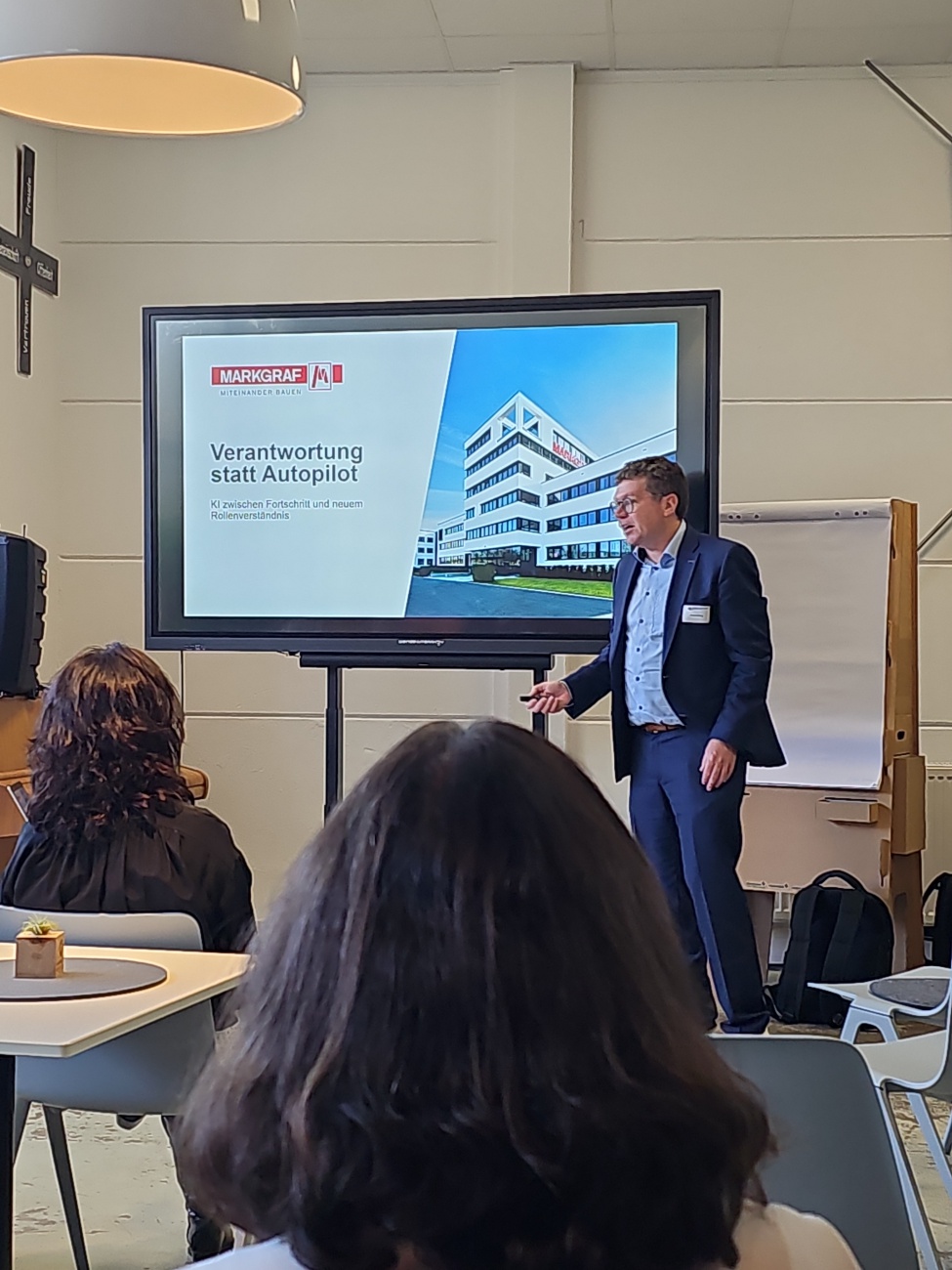 Schon besonders, wie das BetterWorkCamp in Kemnath bei seinem offenen Barcamp-Ansatz für Gesprächsräume statt Rednerbühnen sorgt und so die Frage "Human in the Loop – braucht’s uns noch im Zeitalter der KI?“ mitten in der Nordoberpfalz verhandelt, eine Region, in der Zukunft nicht in Großstädten bleibt, sondern handsam und persönlich gelebt wird. 
Im Bild steht Dr. Stefan König von der W. Markgraf GmbH & Co. KG vor einer Präsentation mit dem Titel "Verantwortung statt Autopilot“, ein Impuls, der sich klar in das zentrale Programm des Tages einfügt. 

Seine Rolle als Sprecher taucht auch im inhaltlichen Ablauf des Camps auf, etwa unter "Verantwortung statt Autopilot“, und macht deutlich, dass es nicht um Technik-, sondern um Steuerungsverantwortung geht. 

Die Szenerie spiegelt die Arbeitsatmosphäre des Camps: 
ein heller, unkomplizierter Raum, in dem Köpfe zusammenkommen, um gemeinsam herauszufinden, welche Rolle der Mensch in der KI-gesteuerten Arbeitswelt künftig übernehmen sollte.

Artikel mit KI-Unterstützung