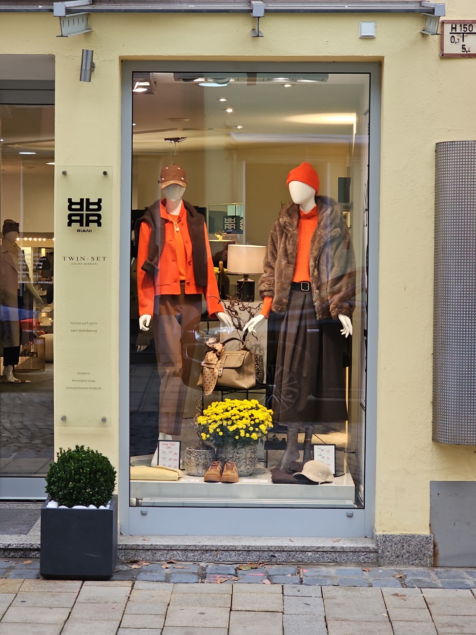 Ein lebendiger Farbpunkt im Schaufenster – inmitten des herbstlichen Grau der Marktredwitzer Innenstadt strahlen zwei stilvoll gekleidete Mannequins in kräftigem Orangeton und beweisen, dass die Saison in feurigen Nuancen aufblüht. 

Das rechte Ensemble kombiniert eine warme Wollmütze und Pullover mit einer weich fallenden Felljacke über einem eleganten, wadenlangen Rock, während links ein passender Blazer mit dunkler Hose samt Halstuch-Herzstück den klassischen Business-Look aufpeppt. Unten im Zentrum sorgt ein üppiger Topf Chrysanthemen in sonnigem Gelb für einen lebendigen Kontrast und einen Hauch Natur. 

Insgesamt vermittelt die Vitrine stilsichere Herbstmode in hochwertig-eleganter Business-Ästhetik – ganz im Sinne der deutschen Premium-Label, wie etwa RIANI, deren Designs opulente Stoffe, raffinierte Schnitte und nachhaltige Materialien vereinen, ohne auf Tragbarkeit zu verzichten (RIANI steht für hochwertige Business-Mode, zeitlose Klassiker und Statement-Pieces)

Artikel erstellt mit KI-Unterstützung