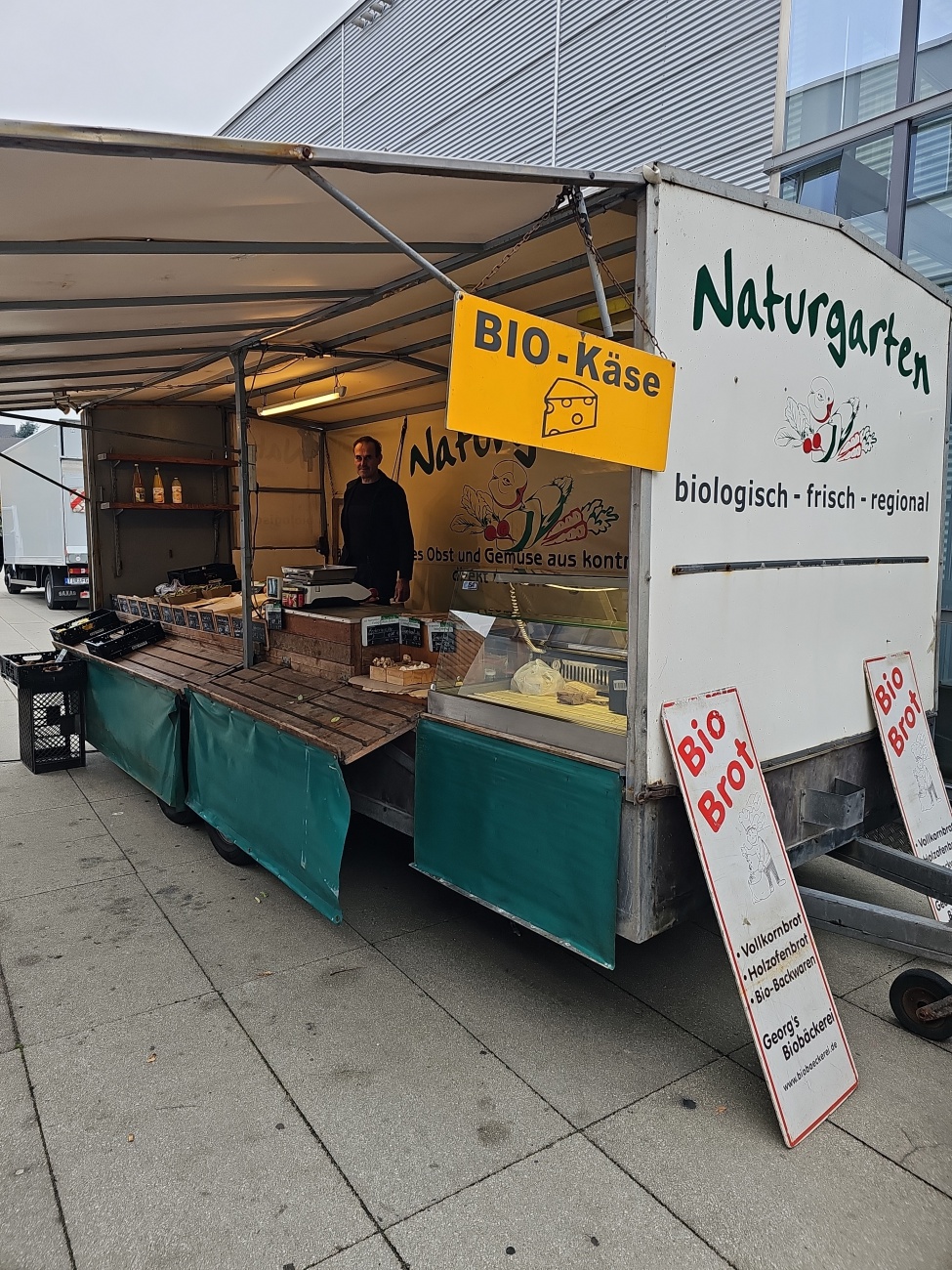 Kurz vorm Feierabend... die Auslagen schon abgeräumt...

Ein grellgelbes Schild mit der Aufschrift "BIO Käse“ hängt über einem liebevoll eingerichteten Marktstand, dessen Rückwand das Logo ..Naturgarten – biologisch -  frisch - regional“ trägt, und vermittelt sofort die Botschaft: 
hier gibt es ehrliche Landwirtschaft zum Anfassen.

Die Theke ist übersichtlich bestückt mit frischen Bio-Backwaren, darunter Vollkorn? und Holzofenbrot, was durch ein Plakat der ..Georgs Biobäckerei“ zusätzlich betont wird – vermutlich ein Lieferant, auch wenn der genaue Herkunftsort unklar bleibt. 
Der Marktstand vermittelt mit seiner rustikalen Holzoptik, den offenen Kisten und der auffälligen Beschilderung ein bodenständiges Flair, das einlädt, im regionalen Angebot zu stöbern und den persönlichen Kontakt mit dem Händler zu suchen. 

Besucherinnen und Besucher des Wochenmarkts auf dem Castelfranco-Emilia-Platz in Marktredwitz dürften hier auf schmackhafte, ökologische Produkte treffen, die handwerkliche Tradition und nachhaltigen Anspruch kombinieren und den regionalen Marktcharakter charmant unterstreichen.