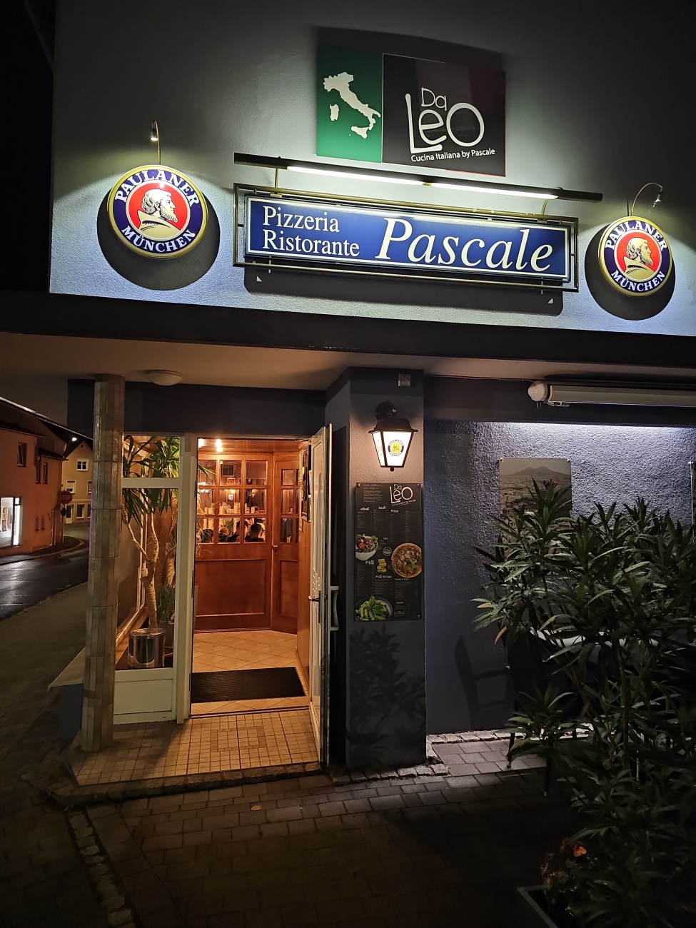 Ein warm beleuchteter Eingang in Pressath, über dem ein Schild „Pizzeria Ristorante Pascale“ mit dem Zusatz „Da Leo“ hängt, lädt mit rustikalen Holztüren und stilvoller Fassadenbeleuchtung zu traditionell italienischer Gastfreundschaft ein.