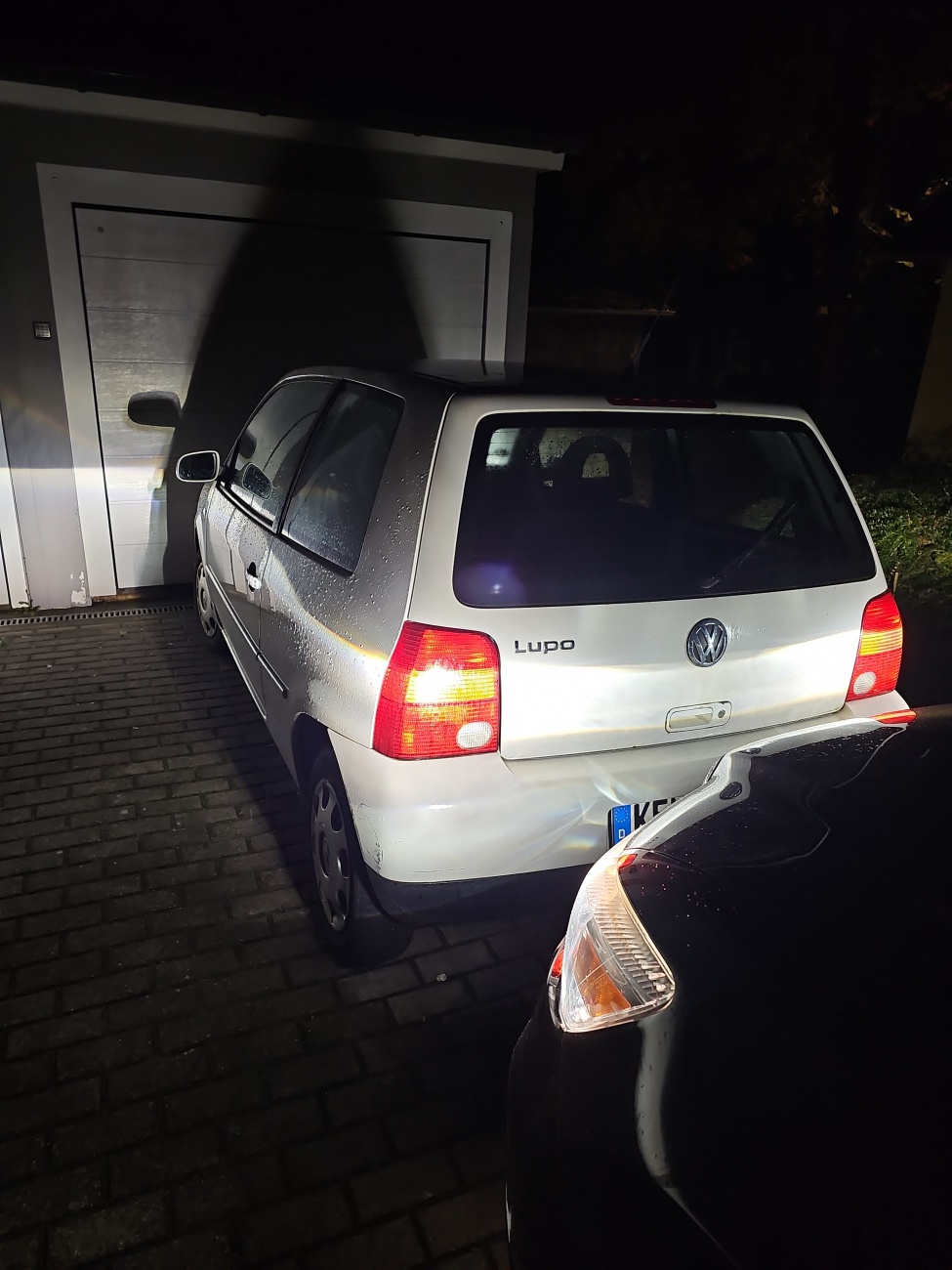 Ein VW Lupo posiert nachts vor der Garage, sein Heck von Regenperlen bedeckt. Der charmante Kleinwagen aus Wolfsburg, einst bekannt als besonders sparsamer 3-Liter-TDI, beweist hier, dass auch Gebrauchsspuren Charakter schaffen.