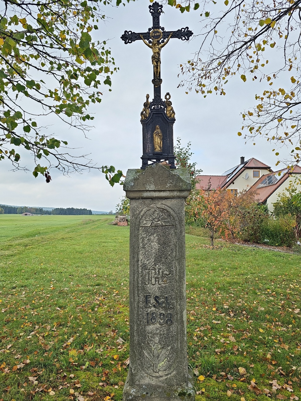 Ein schlichter Bildstock mit eisernem Kruzifix steht am Wegesrand, umgeben von verfärbtem Laub und herbstlichem Himmel. Die vergoldete Christusfigur und das filigrane Marienrelief setzen sich kontrastreich von der verwitterten Steinbasis mit der Jahreszahl 1898 ab.