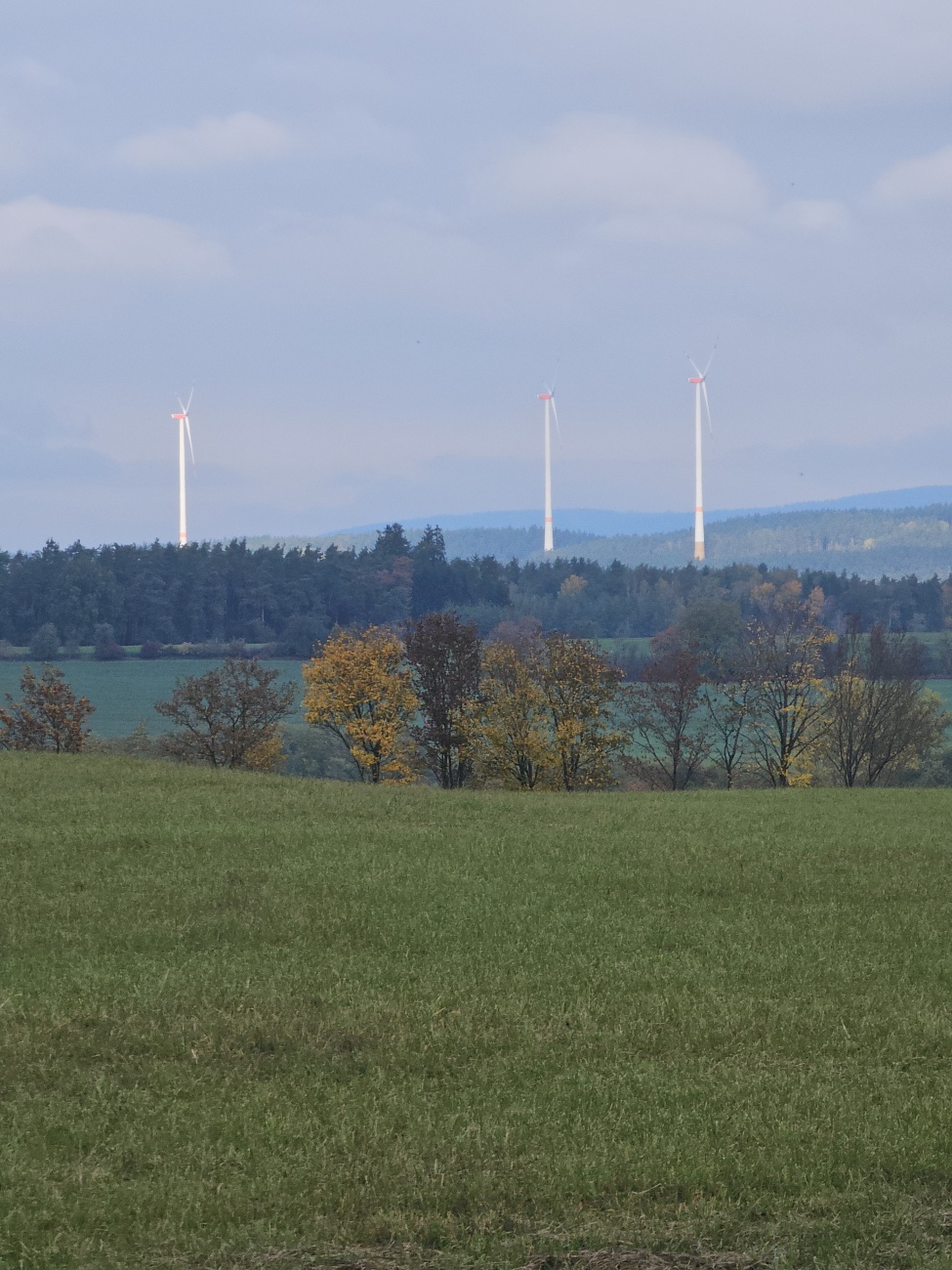 Über herbstlich gefärbten Hügeln stehen mehrere schlanke Windkraftanlagen mit rot markierter Nabe in offener Feldflur, abseits bewohnter Orte. Der Blick reicht über Waldränder bis in die Wälder der Oberpfalz – Sinnbild der regionalen Energiewende.