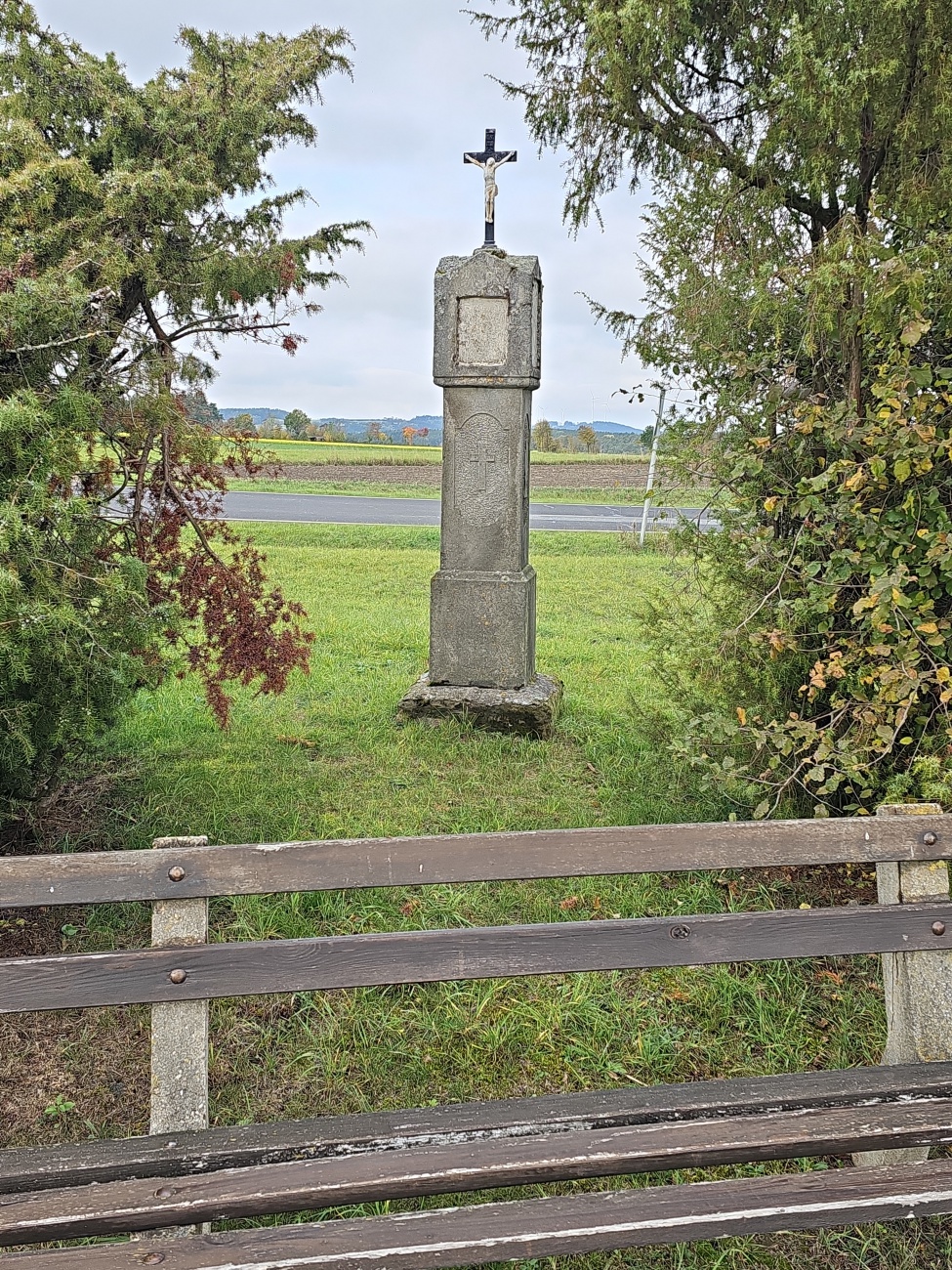 Ein steinernes Wegkreuz mit Korpus Christi steht auf einem etwa 1,5 bis 2 Meter hohen Sockel, umgeben von dichtem Grün und mit Blick über Felder und sanfte Kuppen. Solche Flurkreuze sind typische Kleindenkmäler in ländlichen Gegenden Bayerns.