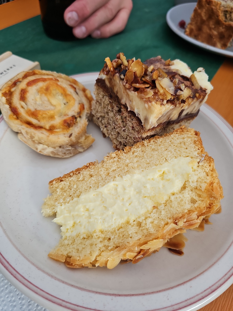 Ein rustikaler Kuchenmix auf schlichtem Teller lädt zum entspannten Kaffeegenuss ein: Helles, buttriges Stück mit Vanillefüllung, ein nussbestreutes Sahnestück und eine spiralförmige, leicht herzhafte Schnecke. Ungekünstelt und bodenständig.