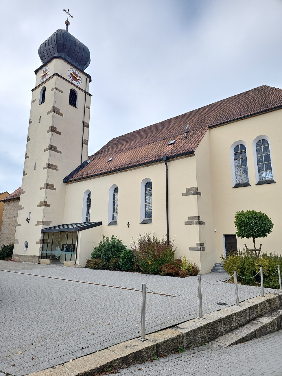 Diese Aufnahme besticht durch den gedrungenen Turm mit markanter Zwiebelhaube, ein typisches Element barocker Kirchen in Bayern. 

Die katholische Pfarrkirche St. Lucia in Schlammersdorf wurde 1775 bis 1777 als Saalkirche mit Satteldach errichtet, das Westlanghaus besteht aus Sandsteinquadern und ist als Baudenkmal eingetragen. 

Besonders ins Auge fällt der spätmittelalterliche Chorturm, um 1500 erbaut und 1684 aufgestockt, dessen charakteristische Haube das Gesamtbild prägt. Amorphe Fenster mit hellen Archivolten gliedern die Wände, deren Fassade in hellem Licht klar wirkt – deutliche Anzeichen für eine Aufnahme bei bedecktem Himmel. 

Im V o r d e r grund lädt ein gepflasterter Platz dazu ein, das Gebäude zu betreten. Die Kirche vereint historische Bauphasen harmonisch, deren Architektur prägt das Ortsbild, zeigt lokale Geschichte und verkörpert barocke Stilvorlieben.