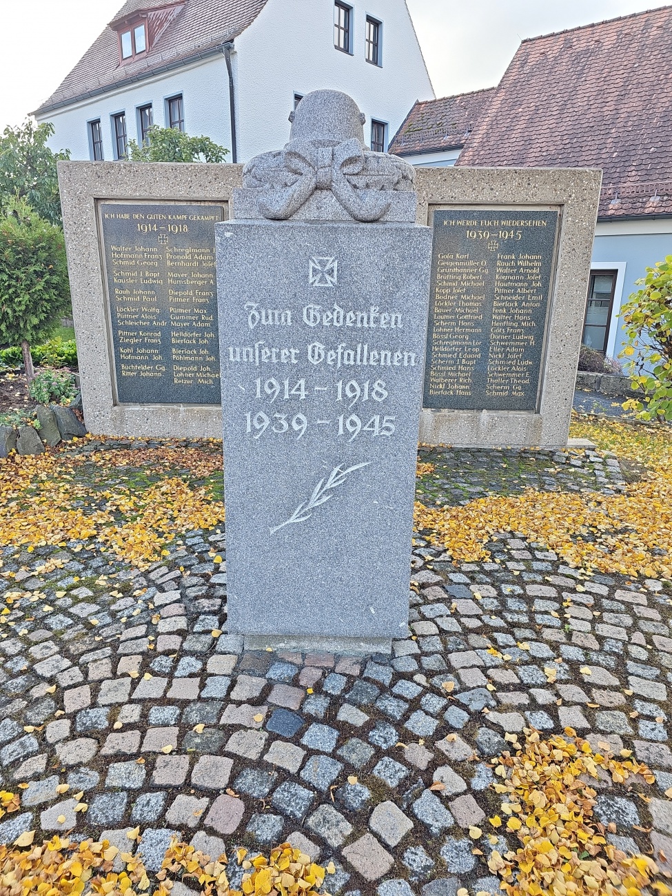 Ein schlicht gehaltener Gedenkstein aus hellem Granit rückt die Namen der Gefallenen zweier Weltkriege in den V o r der grund – eingerahmt von vergoldeter Inschrift, Eichenlaubgravur und dem Eisernen Kreuz. 

Im Herbstlaub vor der Kirche zeichnet das matte Grau des Steins ein würdiges Bild des Erinnerns vor historischer Kulisse. 
Dieses bäuerlich geprägte Mahnmal steht ganz im Zeichen der Trauer um 1914–1918 und 1939–1945, es folgt der üblichen Gestaltungsweise regionaler Kriegerdenkmäler, bei der das Eiserne Kreuz und christliche Symbolik oft zentrale Elemente sind. 

Solche Denkmäler entstanden vielfach auf Initiative von Gemeinden oder Kirchen, um gefallene Männer zu ehren – ohne zugleich patriotische oder triumphale Botschaften zu vermitteln, vielmehr als stille Auflistung der Namen.