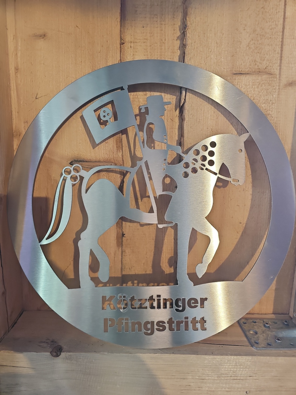 Das Metallrelief mit der Inschrift ..Kötztinger Pfingstritt“ zeigt einen festlich geschmückten Reiter auf einem Pferd und erinnert als stilisiertes Symbol an die traditionsreiche Pferdewallfahrt im Bayerischen Wald. 