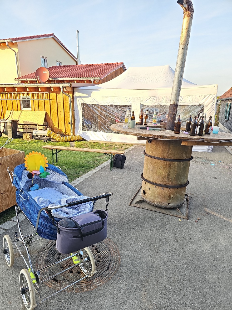 Dorfleben: Im Hof steht ein alter Kinderwagen mit blau-gemusterter Decke und bunten Stofftieren, darunter eine Blumenfigur, neben einer rustikalen Fass-Bar im provisorischen Partyzelt. 

Leere Bierflaschen auf dem Tresen deuten auf ein entspanntes Beisammensein hin.