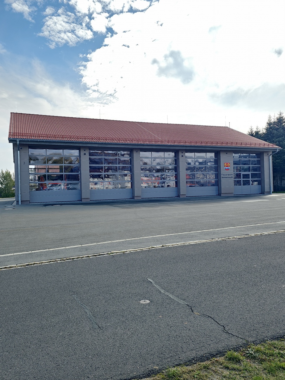 Das moderne Feuerwehrhaus mit seiner klaren Architektur und den großen, verglasten Toren steht am Rand eines weitläufigen Areals – bereit für den schnellen Einsatz, wenn Hilfe gebraucht wird. Die Fassade wirkt durch die Kombination aus funktionalem Grau und roten Akzenten sachlich, aber gepflegt. Das Gebäude spiegelt in den Scheiben die umliegende Landschaft und Fahrzeuge wider, was dem nüchternen Zweckbau eine interessante, fast lebendige Note verleiht.

Feuerwehren in Gemeinden wie Immenreuth übernehmen heute viel mehr als den klassischen Brandschutz: Sie unterstützen bei Unfällen, Unwettern und leisten technische Hilfe – oft ehrenamtlich, rund um die Uhr. Der technisch ausgestattete Standort steht für Sicherheit und Gemeinschaftsgeist, der in ländlichen Regionen besonders wichtig ist. Das Gebäude selbst verrät nicht viel Aufregendes auf den ersten Blick, aber es ist ein stiller Garant dafür, dass im Ernstfall alles bereitsteht.