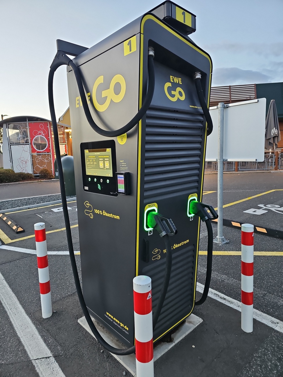 Mitten auf einem Parkplatz steht eine EWE Go-Schnellladestation und verdeutlicht, wie stark sich Mobilität in den letzten Jahren gewandelt hat. Früher dominierten Tankstellen das Bild, heute prägen innovative Ladestationen viele Orte – sichtbar, markant, mit konsequent nachhaltiger Ausrichtung. Die deutliche Beschriftung macht klar, dass hier ausschließlich Ökostrom fließt und setzt damit ein Zeichen für die Verkehrswende.

Gelbe Akzente an der Ladesäule und das große Display laden förmlich zum Laden ein, während rote Poller den Bereich gut absichern. Das Design überzeugt mit einer klaren Linienführung: Sichtbare Kabel, gut erreichbare Ladeanschlüsse und eine benutzerfreundliche Bedienung sorgen für unkomplizierte Nutzung.

Im Hintergrund zeichnen moderne Gebäude und Gastronomie ein lebendiges Bild der Umgebung. Ladepunkte wie dieser sind längst mehr als nur Technik – sie gehören inzwischen zum alltäglichen Stadtbild, gerade in zukunftsorientierten Regionen wie dem Landkreis Neustadt an der Waldnaab.