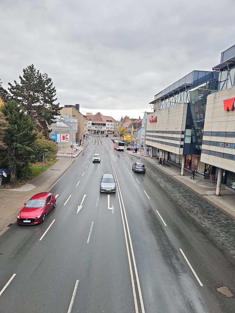 Über einer vielbefahrenen Straße im Zentrum von Bayreuth spannt sich ein grauer Himmel. Links reihen sich Bäume und Geschäftshäuser, rechts dominiert ein Einkaufszentrum mit markanter Glasfassade. Autos, ein Linienbus und wenige Passanten beleben die nasse Szenerie und spiegeln den Wandel der Stadt wider.
