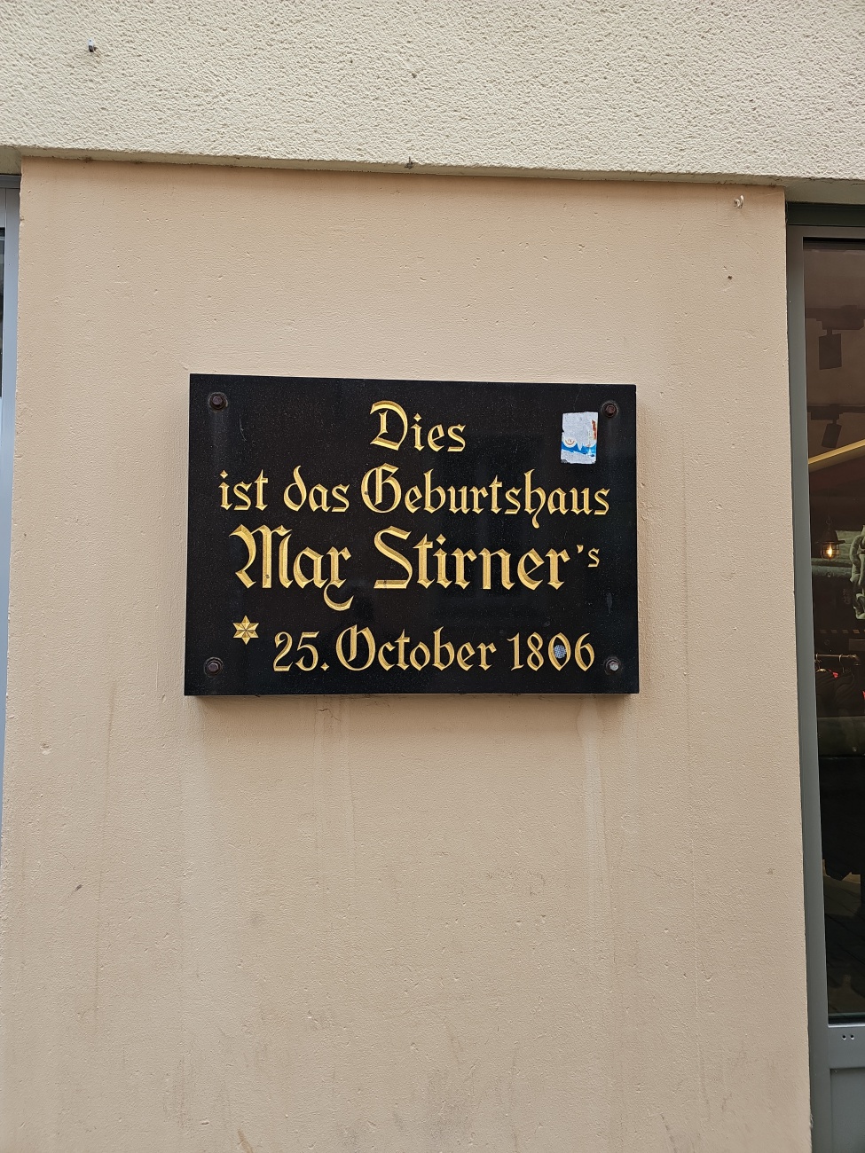 Dezent, fast unscheinbar prangt die schwarze Gedenktafel an der Wand und erinnert an einen prominenten Namen: 
Max Stirner wurde hier geboren, am 25. Oktober 1806.
Hinter diesem Namen verbirgt sich einer der provokantesten Denker des 19. Jahrhunderts, der mit seinen Gedanken über Individualismus und Anarchie weit über Bayreuth hinaus Wirkung entfaltete. 
Sein Hauptwerk "Der Einzige und sein Eigentum" sorgte für kontroverse Diskussionen und verschaffte Stirner bis heute einen festen Platz in der Philosophiegeschichte.

Gerade im Stadtbild geht solche Historie schnell unter, wenn man nicht gezielt hinsieht. 
Die Fassade ist schlicht, kein Prunk, keine Besucherströme – eher fast übersehbar, trotz der goldenen Lettern. Dennoch erhält dieser Ort durch die kleine Platte eine zweite, verborgene Ebene: 

Die Erinnerung bleibt wach, und wer hier vorbeikommt, begegnet unvermittelt einem überraschenden Kapitel Stadtgeschichte.