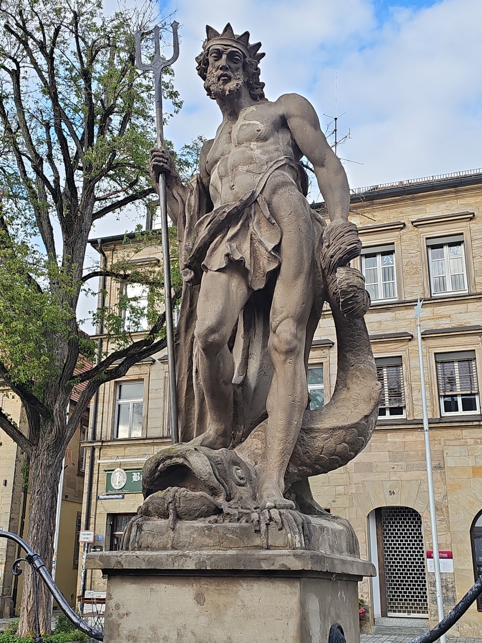 Mit kräftigem Blick, Dreizack und aufgebauschten Wellen als Umhang dominiert der steinerne Neptun diesen Platz in Bayreuth. 

Die Detailverliebtheit an Bart, Haar und Muskulatur zeugt von handwerklichem Können, während der kniende Meeresgott von einem gewundenen Fisch begleitet wird. 

Solche Brunnenfiguren waren in deutschen Residenzstädten einst beliebte Symbole für Wohlstand und für das Zusammenspiel von Mensch und Naturgewalten. Auch in Bayreuth hat die Figur ihre Vergangenheit: 

Der Neptunbrunnen entstand um die Mitte des 18. Jahrhunderts und ist bis heute eines der vertrauten Wahrzeichen der Innenstadt. 
Im Hintergrund deutet das sandfarbene Bürgerhaus auf die historische Bebauung der Umgebung hin, alles in allem wirkt die Szene wie ein Denkmal vergangener Pracht, das bis heute ins Auge fällt.