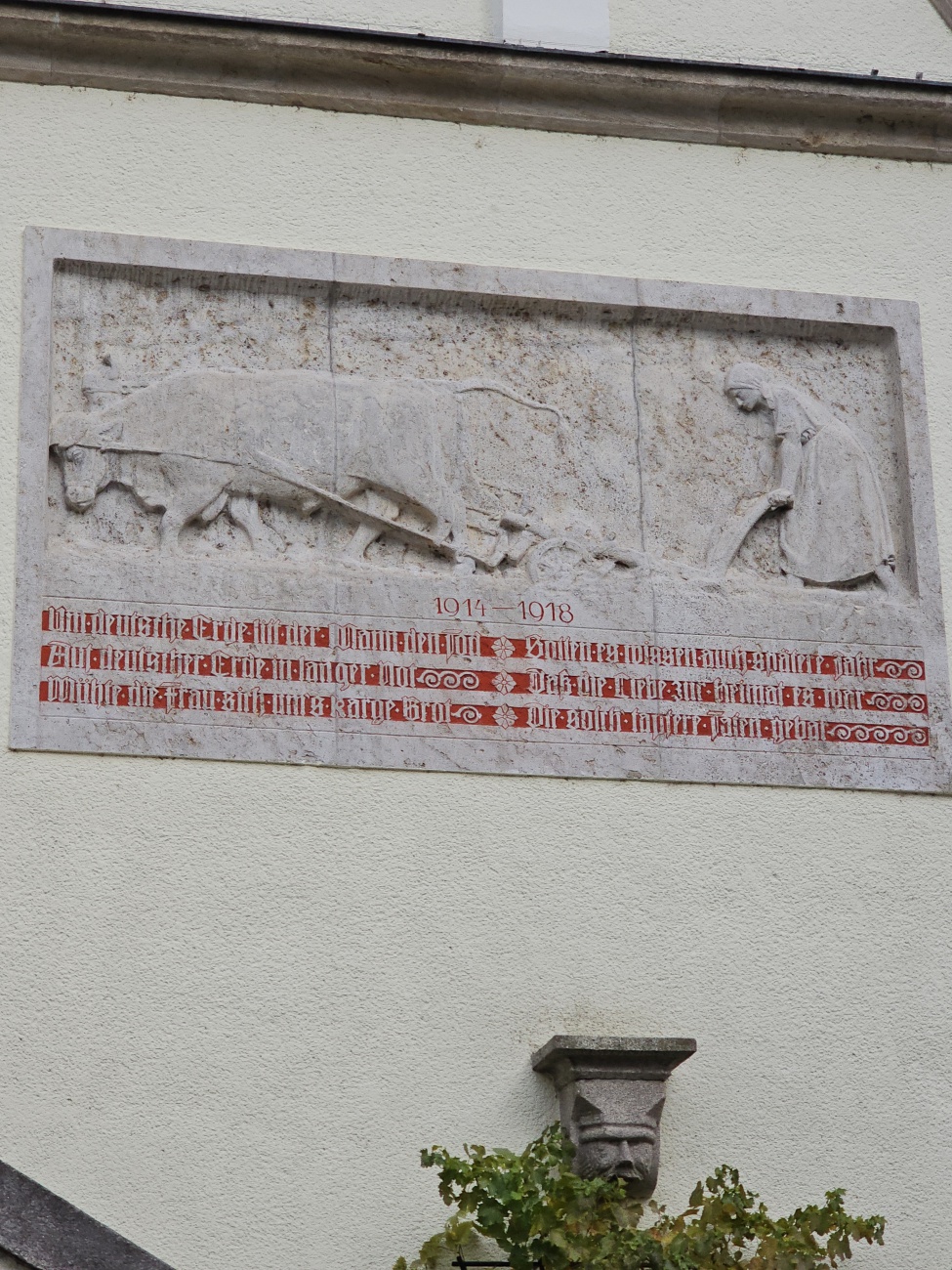 An der Fassade zieht ein Relief aus hellem Stein die Aufmerksamkeit auf sich: Es zeigt eine Frau, die mit einem Ochsengespann ein Feld pflügt – eine eindrucksvolle Szene, die unmittelbar an die Zeit des Ersten Weltkriegs erinnert. In diesen Jahren, als zahlreiche Männer zum Kriegsdienst eingezogen wurden, lag die Verantwortung für die landwirtschaftliche Arbeit und den Fortbestand der Höfe auf den Schultern der Frauen. Die darunter eingravierten Jahreszahlen 1914–1918 und der auffällige rote Schriftzug machen deutlich, dass es sich um ein Mahnmal handelt.

Motivisch ist das Relief typisch für bayerische Kriegerdenkmäler: Neben den gefallenen Soldaten würdigt es auch jene, die zuhause unermüdlich für das Überleben der Familien und Dorfgemeinschaften sorgten. Besonders in ländlichen Gegenden wie Weiden in der Oberpfalz bleibt die enge Verbindung zur Landwirtschaft sichtbar. Das Mahnmal erinnert daran, wie der Krieg die Gemeinschaft tiefgreifend veränderte und den Alltag vieler Menschen von Entbehrung und Pflichtbewusstsein prägte.