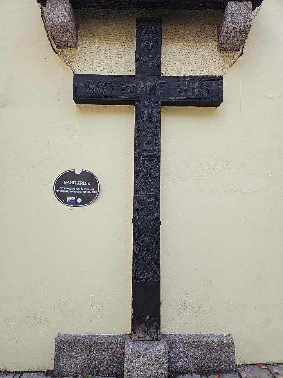 An einer hellen Hauswand in Weiden in der Oberpfalz lehnt ein schwarzes Holzkreuz mit markanten Ritzmustern und der Inschrift „Gott mit uns“. 

Eine Infotafel informiert, dass es sich um ein Nagelkreuz aus der Zeit des Ersten Weltkriegs handelt. Drahtnetz und Steinsockel zeigen, welch bedeutsames Andenken das Kreuz heute noch für die Stadt ist.