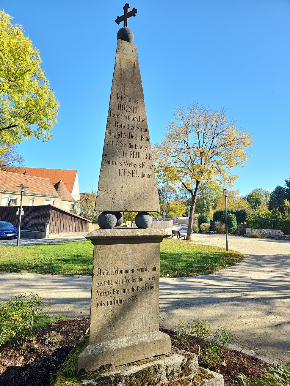 Im gepflegten Grün von Kemnath erhebt sich ein eindrucksvoller Obelisk aus Sandstein, dessen Inschrift an Johann Baptist Hoesel erinnert. Hoesel, geboren 1846, diente als Militärmusiker und verstarb 1870 im jungen Alter. Solche Mahnmale finden sich gerade in kleineren Orten und erzählen leise, aber eindringlich vom Schicksal einzelner Menschen – persönlich und zurückhaltend, fernab der großen städtischen Monumente.

Die Patina des Steins unterstreicht seine Erdverbundenheit und verleiht ihm zusammen mit den klassizistischen Kugeln und dem Kreuz typisch spätklassizistische Züge. Bemerkenswert ist der Zusatz auf der unteren Seite: Das Monument entstand 1873 nach der „Vergößerung dieses Friedhofs“. Hier zeigt sich, wie das Gedenken an das Individuum in der wachsenden Friedhofsanlage einen festen Platz fand.

Unter herbstlichem Laub und blauem Himmel steht der Stein heute fest eingebettet im Alltag, lädt zum Innehalten ein und bleibt dennoch unaufdringlich.