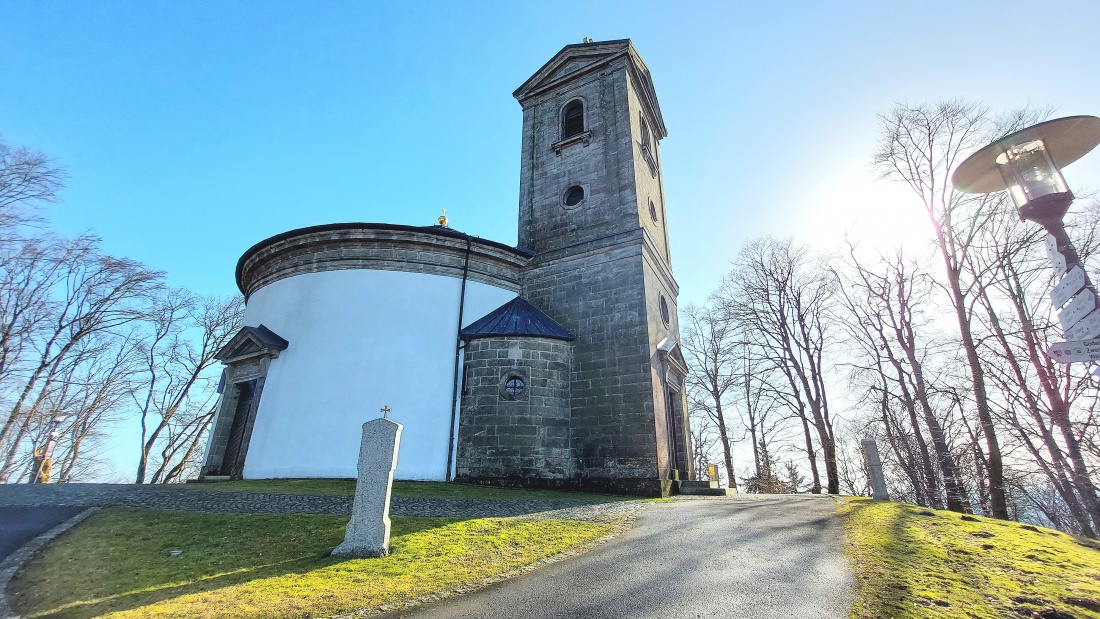 Die Wallfahrtskirche auf dem Armesberg bei schönem Frühlingswetter

Bei schönem Frühlingswetter zeigt sich die Wallfahrtskirche zur Heiligsten Dreifaltigkeit auf dem Armesberg von ihrer eindrucksvollsten Seite. Der Armesberg erhebt sich bis auf 731 Meter über Normalnull und ist ein markanter Basaltkegel im nördlichen Teil der Oberpfalz. Als ehemaliger Vulkanschlot ragt er deutlich aus der umgebenden Landschaft hervor und bietet eine weite Aussicht über das Fichtelgebirge und das Umland.

Auf seinem Gipfel steht die Kirche mit ihrem charakteristischen runden Baukörper. Die hell verputzten Mauern leuchten im Sonnenlicht, während sich darüber der kleine Turm mit Kreuz gegen den blauen Himmel abzeichnet. Das schlichte, aber kraftvolle Erscheinungsbild des Gotteshauses fügt sich harmonisch in die natürliche Umgebung ein. Gerade im Frühling, wenn die Bäume austreiben und die Wiesen wieder grün werden, entsteht eine besonders freundliche und zugleich feierliche Atmosphäre.

Die Geschichte des Wallfahrtsortes reicht bis ins 17. Jahrhundert zurück. In den Jahren 1677 und 1678 wurde auf dem Berg eine erste Kapelle errichtet. Die heutige Kirche entstand später. Der Bau begann im Jahr 1822 und die Weihe erfolgte 1836. Seitdem ist der Armesberg ein bedeutender Ort des Glaubens und der regionalen Wallfahrt. Ein Kreuzweg führt hinauf zur Kirche und unterstreicht die religiöse Bedeutung des Berges.

Wer an einem klaren Frühlingstag den Gipfel erreicht, spürt die besondere Verbindung von Natur, Geschichte und Spiritualität. Das Licht, die frische Luft und die weite Aussicht verleihen dem Ort eine ruhige und zugleich erhabene Stimmung. Die Kirche auf dem Armesberg ist damit nicht nur ein historisches Bauwerk, sondern auch ein lebendiger Treffpunkt für Pilger, Wanderer und Besucher, die hier oben einen Moment der Stille suchen.