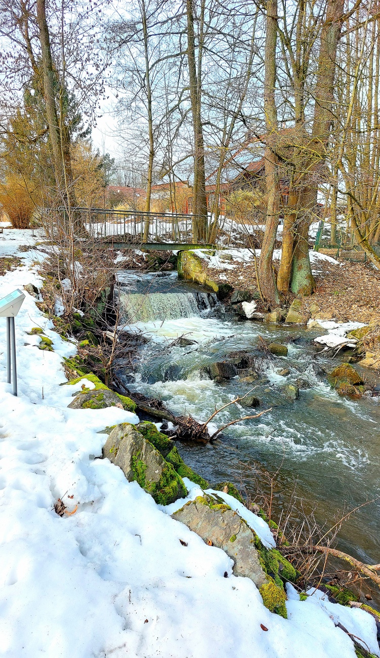 Ein Wasserfall bzw. Wehr in Kemnath...

Das Bild zeigt einen idyllischen Bachlauf in Kemnath, eingebettet in eine ruhige Winterlandschaft.

Schmelzwasser rauscht über kleine Felsen und bildet einen natürlichen Wasserfall unter einer schmalen Brücke.

Schnee, Moos und kahle Bäume verleihen der Szene eine besondere, fast märchenhafte Stimmung.

Die Verbindung aus fließendem Wasser und winterlicher Natur strahlt Ruhe und Frische aus.
Dieser Ort lädt zum Innehalten ein und zeigt die natürliche Schönheit der Region Kemnath.