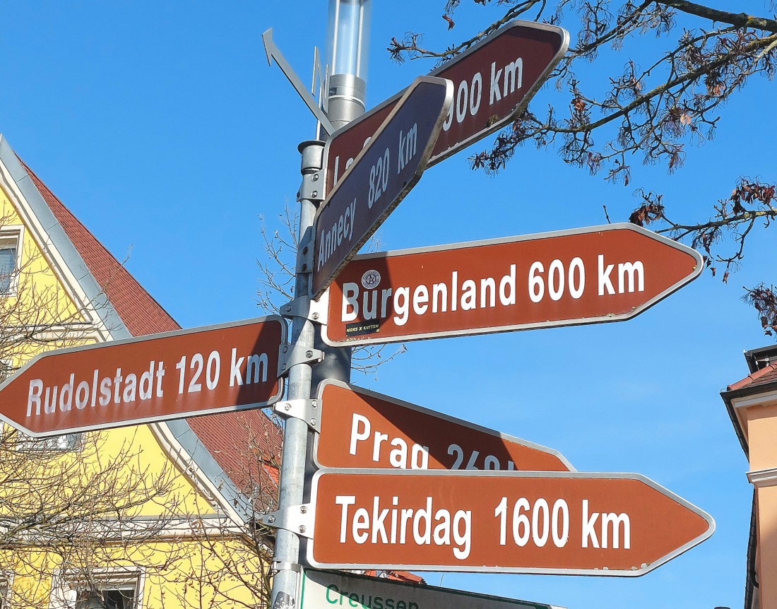 Alle Wege... aus Bayreuth raus...

Wege die verbinden

Dieses Foto zeigt mehr als nur Wegweiser. Es erzählt von Sehnsucht, Fernweh und grenzenloser Freiheit. 

Die Schilder weisen in unterschiedliche Richtungen nach Rudolstadt, Burgenland, Prag, Tekirdag, und sogar bis nach Almere. Jede Richtung steht für eine eigene Geschichte, eine andere Kultur und neue Begegnungen.

Unter dem strahlend blauen Himmel wirken die Entfernungsangaben wie Einladungen. 120 Kilometer, 600 Kilometer oder 1600 Kilometer sind keine bloßen Zahlen, sondern Inspiration. Sie erinnern uns daran, dass die Welt offen ist und jeder Weg mit einem ersten Schritt beginnt.

Zwischen traditioneller Architektur und kahlen Ästen im V o r d e r grund entsteht eine besondere Atmosphäre. Verwurzelt im Hier und Jetzt und zugleich mit Blick in die Ferne. Genau darum geht es. Die Verbindung von Heimat und Horizont.

Dieses Bild steht für Orientierung, für Entscheidungen und für den Mut neue Wege zu gehen. Denn egal wohin die Reise führt, jeder Weg beginnt genau hier.