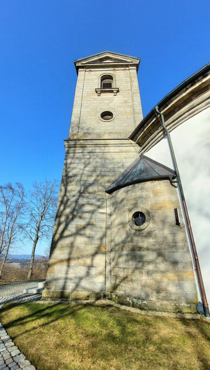 Die Wallfahrtskirche auf dem Armesberg bei schönem Frühlingswetter

Bei schönem Frühlingswetter zeigt sich die Wallfahrtskirche zur Heiligsten Dreifaltigkeit auf dem Armesberg von ihrer eindrucksvollsten Seite. Der Armesberg erhebt sich bis auf 731 Meter über Normalnull und ist ein markanter Basaltkegel im nördlichen Teil der Oberpfalz. Als ehemaliger Vulkanschlot ragt er deutlich aus der umgebenden Landschaft hervor und bietet eine weite Aussicht über das Fichtelgebirge und das Umland.

Auf seinem Gipfel steht die Kirche mit ihrem charakteristischen runden Baukörper. Die hell verputzten Mauern leuchten im Sonnenlicht, während sich darüber der kleine Turm mit Kreuz gegen den blauen Himmel abzeichnet. Das schlichte, aber kraftvolle Erscheinungsbild des Gotteshauses fügt sich harmonisch in die natürliche Umgebung ein. Gerade im Frühling, wenn die Bäume austreiben und die Wiesen wieder grün werden, entsteht eine besonders freundliche und zugleich feierliche Atmosphäre.

Die Geschichte des Wallfahrtsortes reicht bis ins 17. Jahrhundert zurück. In den Jahren 1677 und 1678 wurde auf dem Berg eine erste Kapelle errichtet. Die heutige Kirche entstand später. Der Bau begann im Jahr 1822 und die Weihe erfolgte 1836. Seitdem ist der Armesberg ein bedeutender Ort des Glaubens und der regionalen Wallfahrt. Ein Kreuzweg führt hinauf zur Kirche und unterstreicht die religiöse Bedeutung des Berges.

Wer an einem klaren Frühlingstag den Gipfel erreicht, spürt die besondere Verbindung von Natur, Geschichte und Spiritualität. Das Licht, die frische Luft und die weite Aussicht verleihen dem Ort eine ruhige und zugleich erhabene Stimmung. Die Kirche auf dem Armesberg ist damit nicht nur ein historisches Bauwerk, sondern auch ein lebendiger Treffpunkt für Pilger, Wanderer und Besucher, die hier oben einen Moment der Stille suchen.