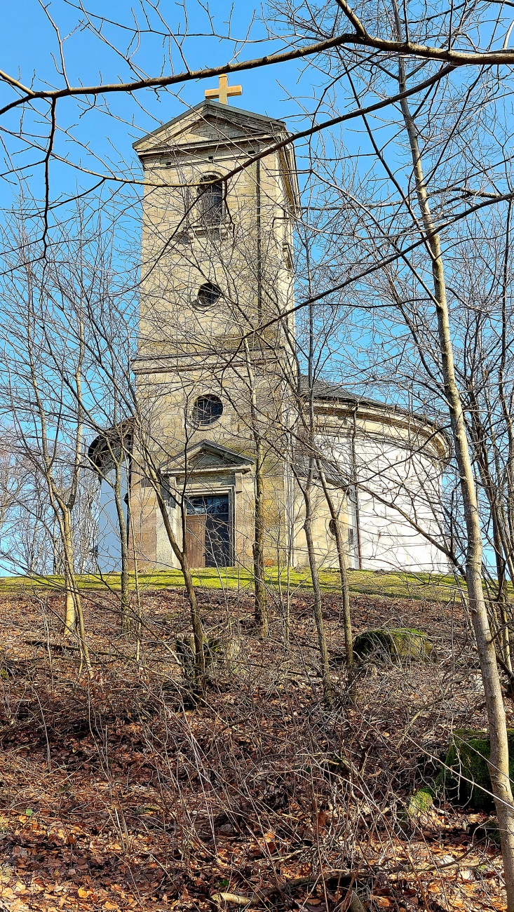 Die Wallfahrtskirche auf dem Armesberg bei schönem Frühlingswetter

Bei schönem Frühlingswetter zeigt sich die Wallfahrtskirche zur Heiligsten Dreifaltigkeit auf dem Armesberg von ihrer eindrucksvollsten Seite. Der Armesberg erhebt sich bis auf 731 Meter über Normalnull und ist ein markanter Basaltkegel im nördlichen Teil der Oberpfalz. Als ehemaliger Vulkanschlot ragt er deutlich aus der umgebenden Landschaft hervor und bietet eine weite Aussicht über das Fichtelgebirge und das Umland.

Auf seinem Gipfel steht die Kirche mit ihrem charakteristischen runden Baukörper. Die hell verputzten Mauern leuchten im Sonnenlicht, während sich darüber der kleine Turm mit Kreuz gegen den blauen Himmel abzeichnet. Das schlichte, aber kraftvolle Erscheinungsbild des Gotteshauses fügt sich harmonisch in die natürliche Umgebung ein. Gerade im Frühling, wenn die Bäume austreiben und die Wiesen wieder grün werden, entsteht eine besonders freundliche und zugleich feierliche Atmosphäre.

Die Geschichte des Wallfahrtsortes reicht bis ins 17. Jahrhundert zurück. In den Jahren 1677 und 1678 wurde auf dem Berg eine erste Kapelle errichtet. Die heutige Kirche entstand später. Der Bau begann im Jahr 1822 und die Weihe erfolgte 1836. Seitdem ist der Armesberg ein bedeutender Ort des Glaubens und der regionalen Wallfahrt. Ein Kreuzweg führt hinauf zur Kirche und unterstreicht die religiöse Bedeutung des Berges.

Wer an einem klaren Frühlingstag den Gipfel erreicht, spürt die besondere Verbindung von Natur, Geschichte und Spiritualität. Das Licht, die frische Luft und die weite Aussicht verleihen dem Ort eine ruhige und zugleich erhabene Stimmung. Die Kirche auf dem Armesberg ist damit nicht nur ein historisches Bauwerk, sondern auch ein lebendiger Treffpunkt für Pilger, Wanderer und Besucher, die hier oben einen Moment der Stille suchen.